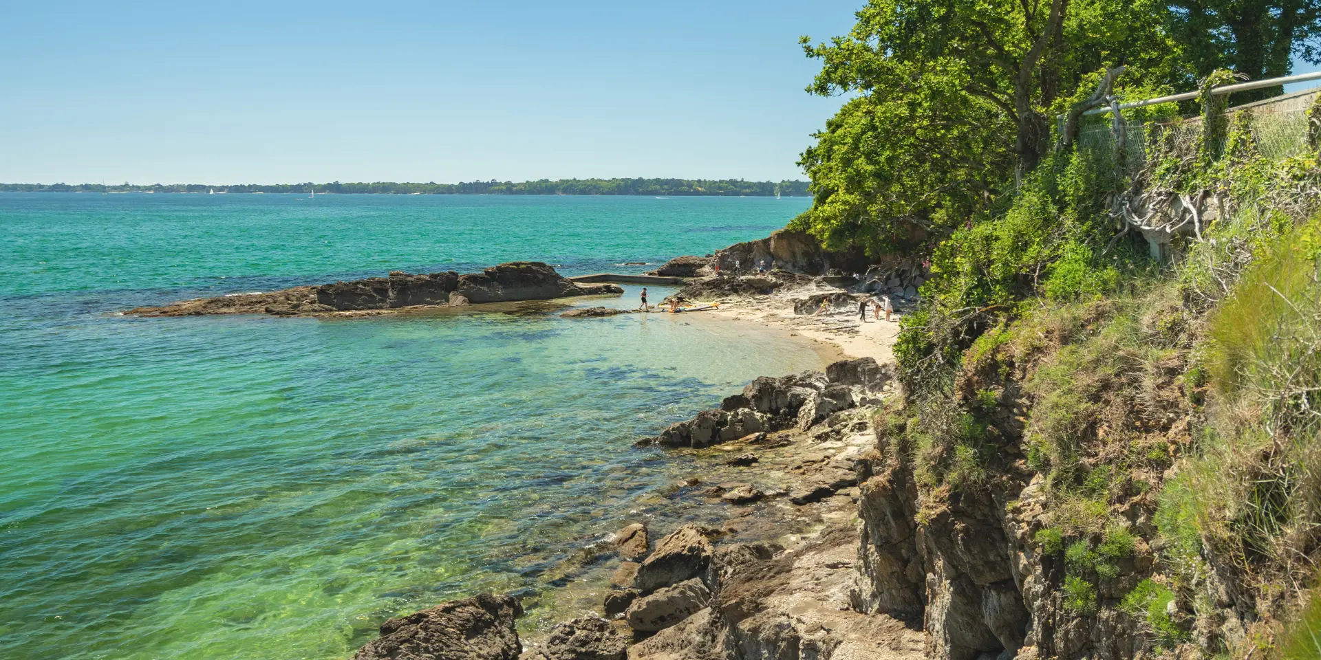 Photo of a rocky coastline in the foreground with transparent turquoise-green water, a small beach in the middle, and on the right a cliff covered with grass and trees under a blue sky.
