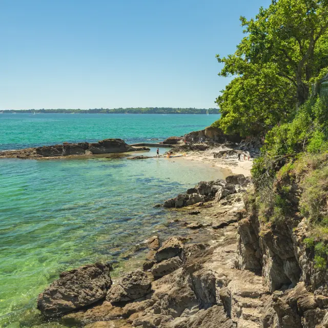 Photo of a rocky coastline in the foreground with transparent turquoise-green water, a small beach in the middle, and on the right a cliff covered with grass and trees under a blue sky.