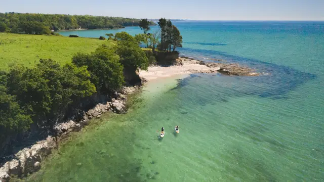 Dieses Foto zeigt eine Küste aus der Vogelperspektive: türkisfarbenes und tiefblaues Wasser, ein kleiner, heller Strand und Felsen, mit einem grünen Landstreifen und Bäumen. Zwei Personen sind mit Kajaks und Paddelbooten in der Nähe der unteren Mitte des Bildes auf dem Wasser unterwegs. Der Himmel ist klar.
