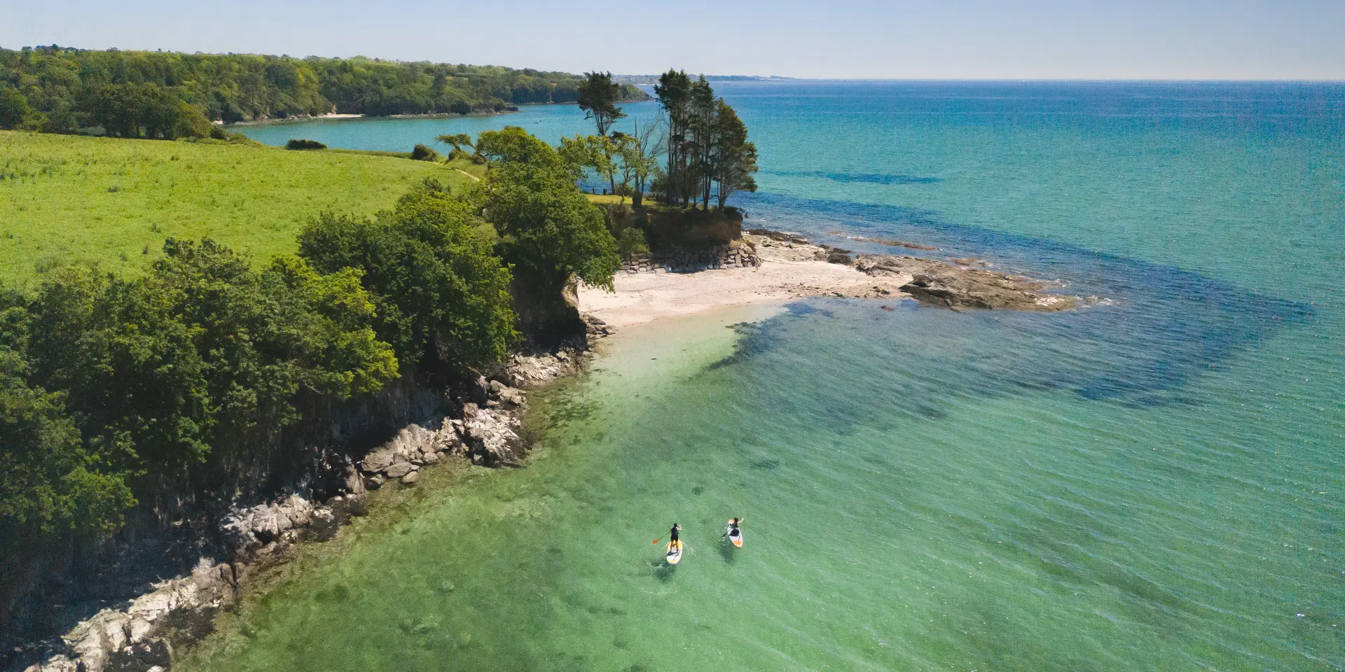 Dieses Foto zeigt eine Küste aus der Vogelperspektive: türkisfarbenes und tiefblaues Wasser, ein kleiner, heller Strand und Felsen, mit einem grünen Landstreifen und Bäumen. Zwei Personen sind mit Kajaks und Paddelbooten in der Nähe der unteren Mitte des Bildes auf dem Wasser unterwegs. Der Himmel ist klar.