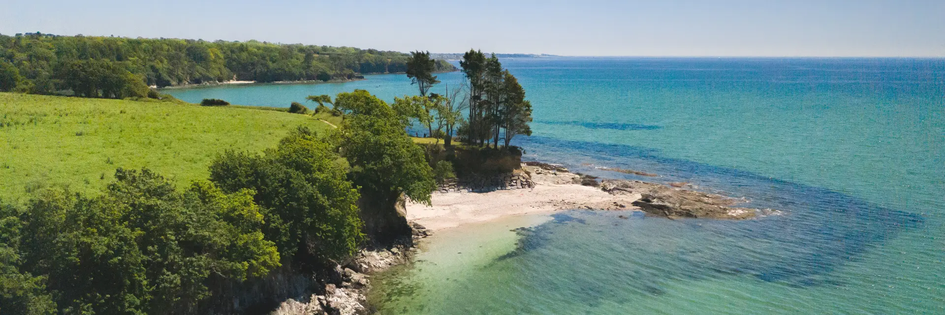 Foto van een luchtfoto van een kustlijn: diepblauw turquoise water, klein helder strand en rotsen, met een overhang van groen land en bomen. Twee mensen zijn op het water in kajaks en paddleboards in het midden onderaan de foto. De lucht is helder.