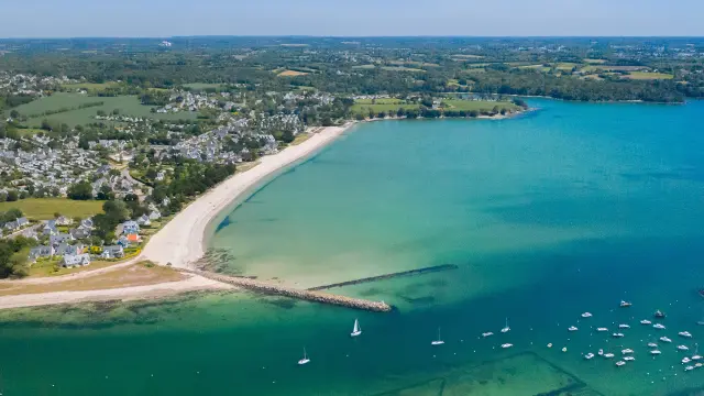 grande photo aérienne d’une plage en arc de cercle et d’une baie à l’eau turquoise. La plage est claire (sable blanc) avec une zone habitée et des champs à l’arrière. Dans l’eau, de nombreux petits bateaux sont mouillés; deux digues/jetées s’avancent dans la mer. En bas de l’image, on distingue des parcs à coquillages sous l’eau.