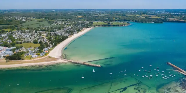 große Luftaufnahme eines bogenförmigen Strandes und einer Bucht mit türkisfarbenem Wasser. Der Strand ist hell (weißer Sand), mit einem bewohnten Gebiet und Feldern dahinter. Im Wasser liegen viele kleine Boote vor Anker; zwei Deiche/Jeeps ragen in das Meer hinein. Im unteren Teil des Bildes sind unter Wasser Muschelparks zu erkennen.