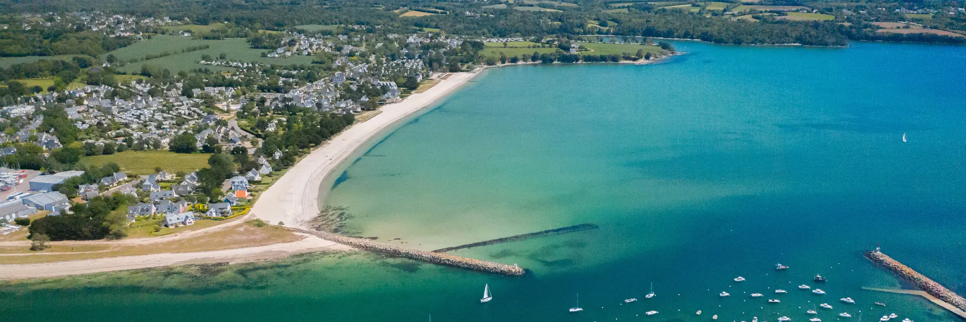 grande photo aérienne d’une plage en arc de cercle et d’une baie à l’eau turquoise. La plage est claire (sable blanc) avec une zone habitée et des champs à l’arrière. Dans l’eau, de nombreux petits bateaux sont mouillés; deux digues/jetées s’avancent dans la mer. En bas de l’image, on distingue des parcs à coquillages sous l’eau.