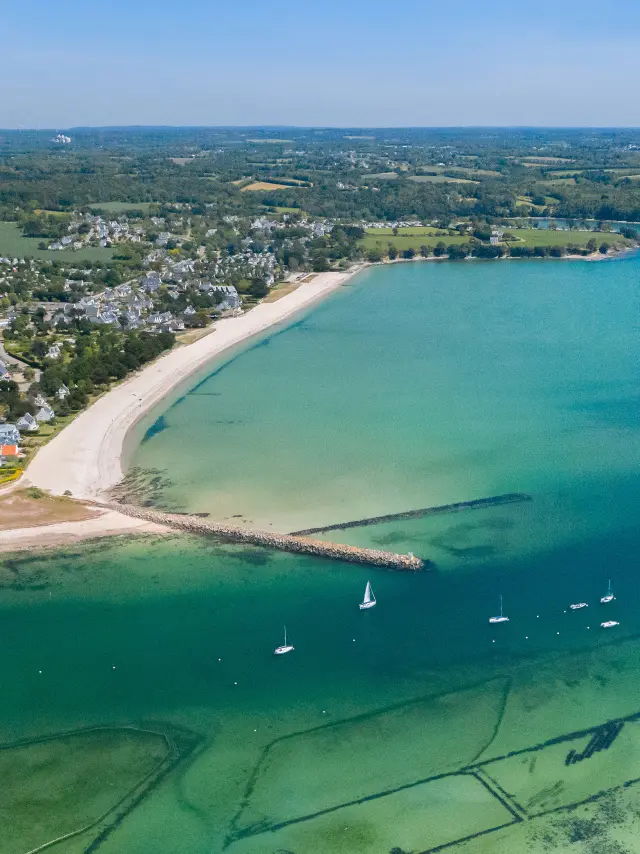 Large aerial photo of an arched beach and a bay with turquoise water. The beach is clear (white sand) with an inhabited area and fields behind. In the water, a number of small boats are moored; two dykes/jetty jut out into the sea. At the bottom of the image, you can see shellfish beds underwater.