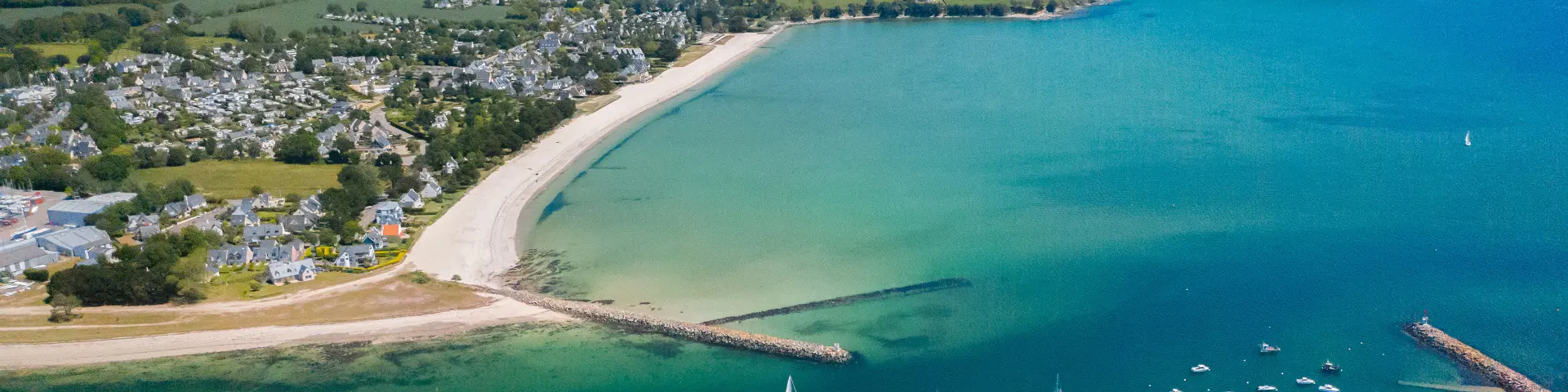Large aerial photo of an arched beach and a bay with turquoise water. The beach is clear (white sand) with an inhabited area and fields behind. In the water, a number of small boats are moored; two dykes/jetty jut out into the sea. At the bottom of the image, you can see shellfish beds underwater.