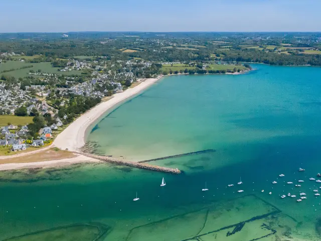 große Luftaufnahme eines bogenförmigen Strandes und einer Bucht mit türkisfarbenem Wasser. Der Strand ist hell (weißer Sand), mit einem bewohnten Gebiet und Feldern dahinter. Im Wasser liegen viele kleine Boote vor Anker; zwei Deiche/Jeeps ragen in das Meer hinein. Im unteren Teil des Bildes sind unter Wasser Muschelparks zu erkennen.