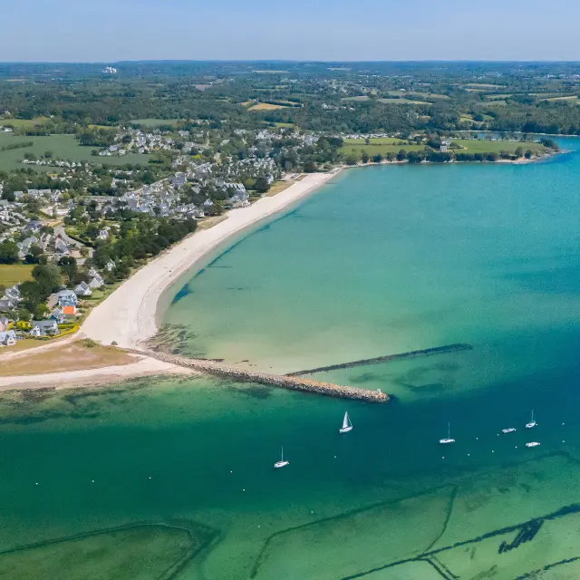 große Luftaufnahme eines bogenförmigen Strandes und einer Bucht mit türkisfarbenem Wasser. Der Strand ist hell (weißer Sand), mit einem bewohnten Gebiet und Feldern dahinter. Im Wasser liegen viele kleine Boote vor Anker; zwei Deiche/Jeeps ragen in das Meer hinein. Im unteren Teil des Bildes sind unter Wasser Muschelparks zu erkennen.