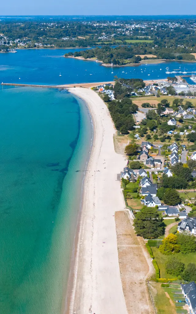 Une photo aérienne d’un littoral. Une longue plage claire borde une mer turquoise. À droite de la plage, une zone résidentielle avec des maisons et des rues. Plus loin, un grand port de plaisance rempli de bateaux, et au fond une anse. Horizon et ciel bleu.