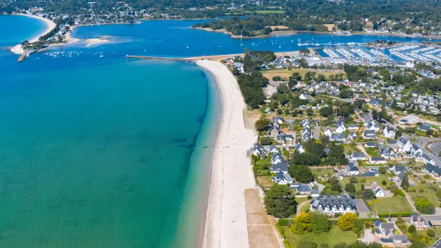 Een luchtfoto van een kustlijn. Een lang, helder strand grenst aan een turquoise zee. Rechts van het strand een woonwijk met huizen en straten. Verderop een grote jachthaven vol boten en een baai op de achtergrond. Horizon en blauwe lucht.