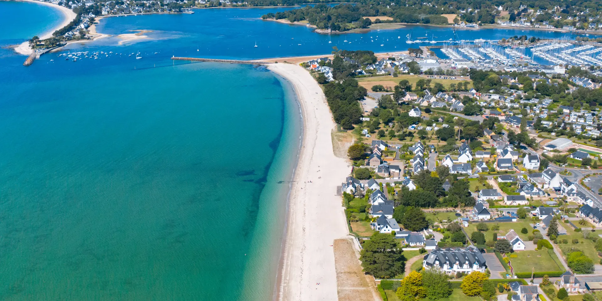 Ein Luftbild einer Küste. Ein langer, heller Strand grenzt an ein türkisfarbenes Meer. Rechts vom Strand befindet sich ein Wohngebiet mit Häusern und Straßen. Weiter hinten ein großer Yachthafen mit vielen Booten und im Hintergrund eine Bucht. Horizont und blauer Himmel.