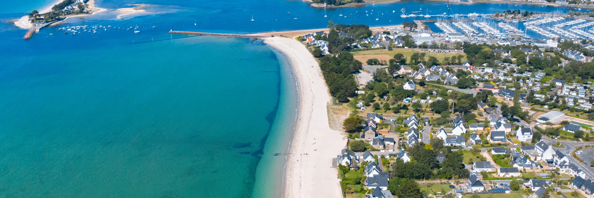 Een luchtfoto van een kustlijn. Een lang, helder strand grenst aan een turquoise zee. Rechts van het strand een woonwijk met huizen en straten. Verderop een grote jachthaven vol boten en een baai op de achtergrond. Horizon en blauwe lucht.