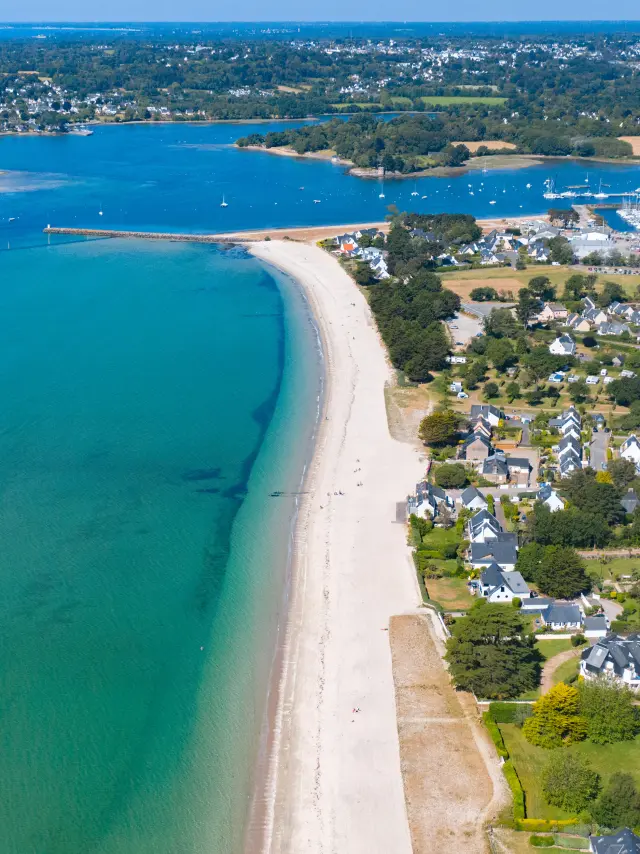 Une photo aérienne d’un littoral. Une longue plage claire borde une mer turquoise. À droite de la plage, une zone résidentielle avec des maisons et des rues. Plus loin, un grand port de plaisance rempli de bateaux, et au fond une anse. Horizon et ciel bleu.