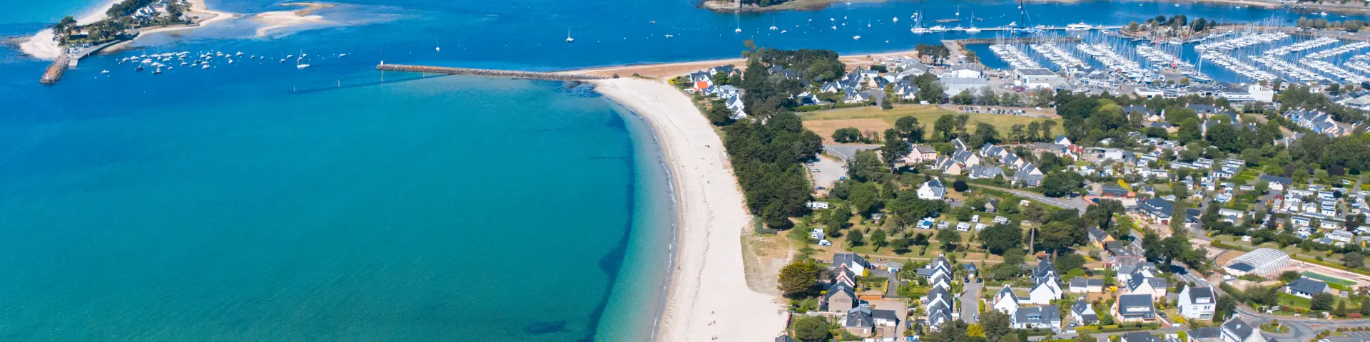 Une photo aérienne d’un littoral. Une longue plage claire borde une mer turquoise. À droite de la plage, une zone résidentielle avec des maisons et des rues. Plus loin, un grand port de plaisance rempli de bateaux, et au fond une anse. Horizon et ciel bleu.
