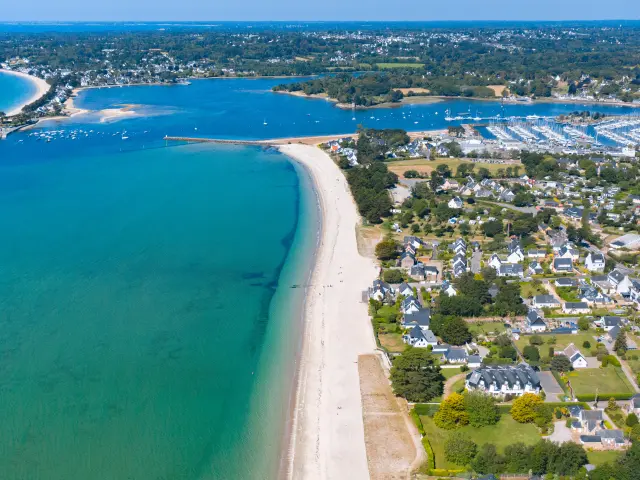 Een luchtfoto van een kustlijn. Een lang, helder strand grenst aan een turquoise zee. Rechts van het strand een woonwijk met huizen en straten. Verderop een grote jachthaven vol boten en een baai op de achtergrond. Horizon en blauwe lucht.