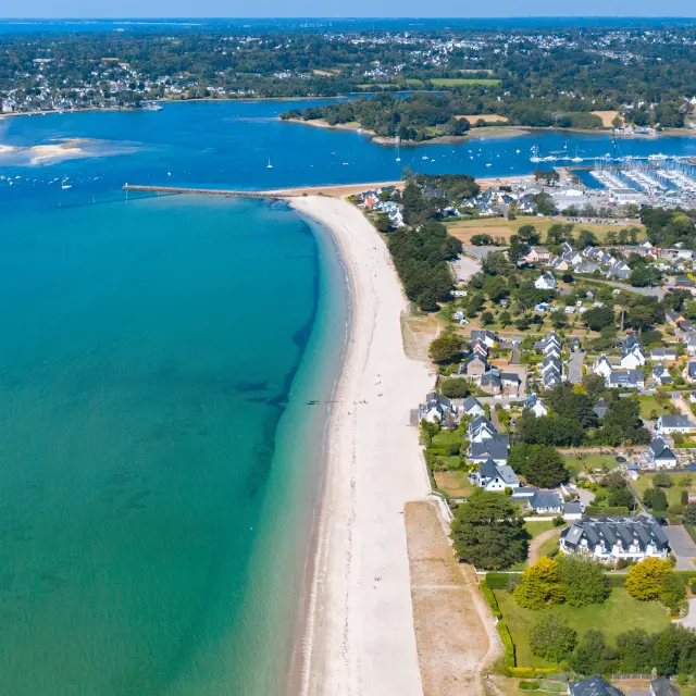 Une photo aérienne d’un littoral. Une longue plage claire borde une mer turquoise. À droite de la plage, une zone résidentielle avec des maisons et des rues. Plus loin, un grand port de plaisance rempli de bateaux, et au fond une anse. Horizon et ciel bleu.