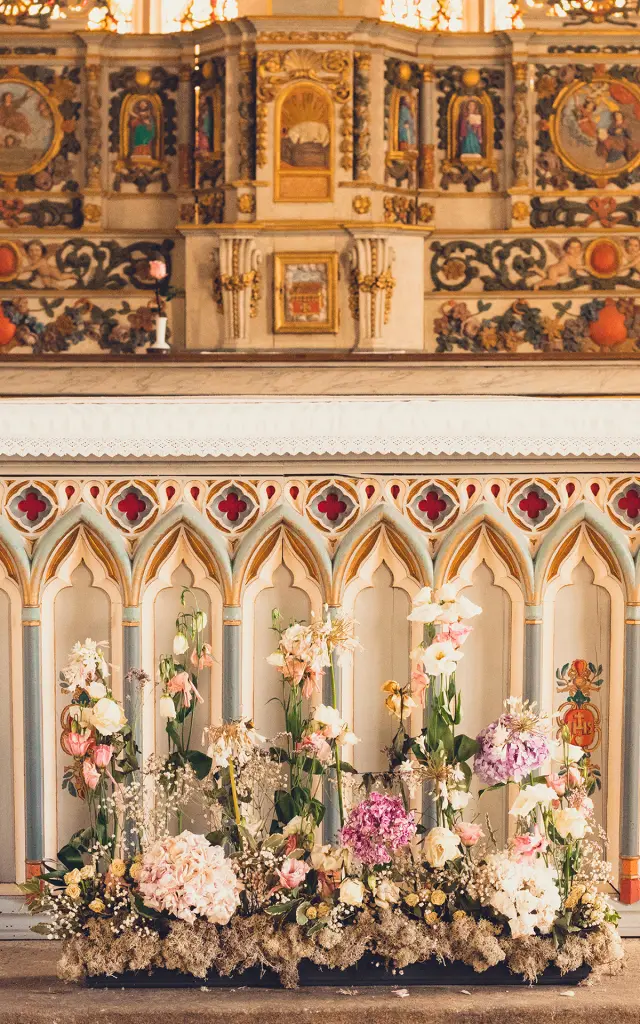 Deze foto toont een rijkelijk versierd kerkaltaar, met lambrisering in gotische stijl en een boeket bloemen aan de voet van het altaar. Daarachter een kleurrijk altaarstuk met verguldsel en decoratieve panelen. De vloer is gemaakt van flagstones en het altaar is verhoogd met treden.