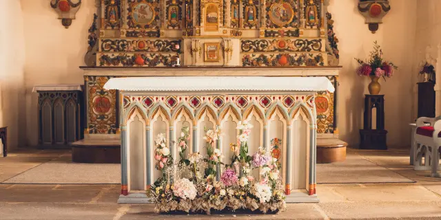 Ein Foto, das einen reich verzierten Kirchenaltar mit einer Verblendung mit gotischen Motiven und einem Blumenstrauß an der Unterseite des Altars zeigt. Dahinter befindet sich ein farbenfroher Altaraufsatz mit Vergoldungen und dekorativen Paneelen. Der Boden besteht aus Steinplatten und der Altar ist durch Stufen erhöht.