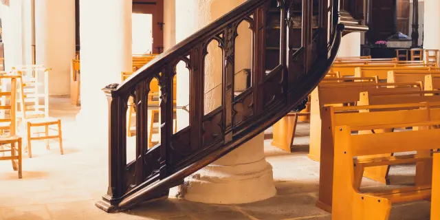 Une photo qui montre l’intérieur d’une église, avec une chaire/rampe d’escalier en bois sombre au premier plan, des colonnes blanches et des rangées de bancs en bois clair à droite, éclairées par une lumière chaude.