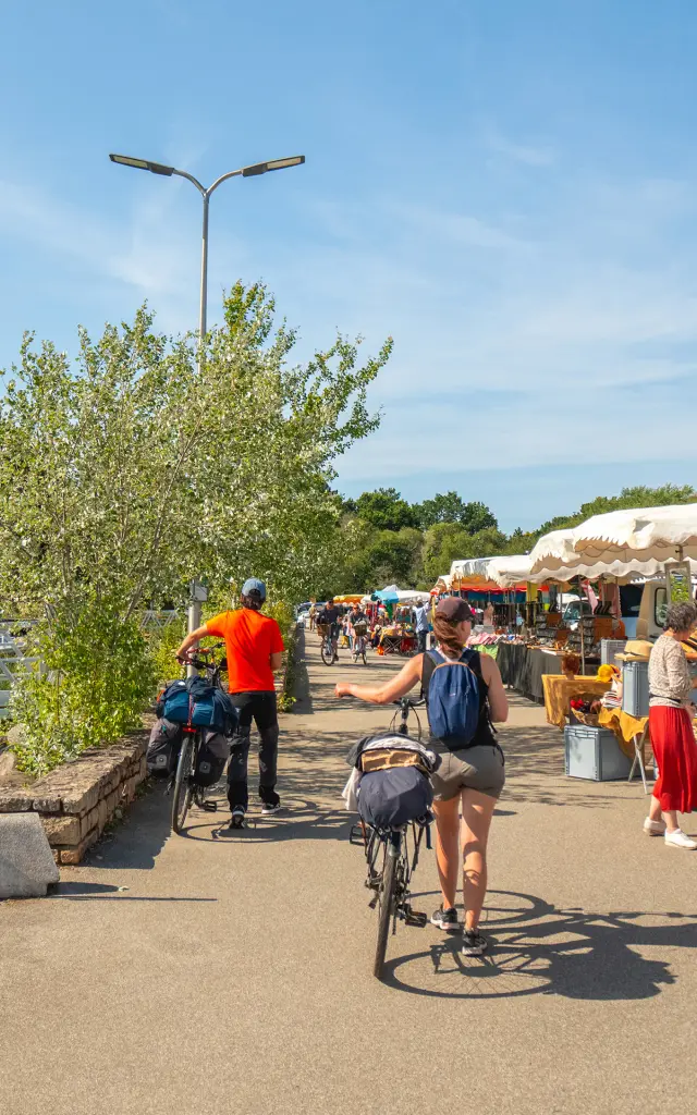 Une photo qui représente un marché en plein air le long d’un port, avec stands sous tentes blanches à droite, des personnes qui marchent, une personne à vélo au centre, et des bateaux/mâts à gauche près de l’eau, sous un ciel bleu.
