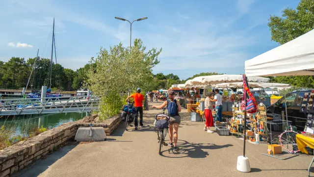 Une photo qui représente un marché en plein air le long d’un port, avec stands sous tentes blanches à droite, des personnes qui marchent, une personne à vélo au centre, et des bateaux/mâts à gauche près de l’eau, sous un ciel bleu.