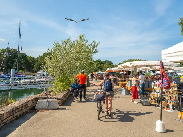 A photo showing an open-air market along a harbour, with white tented stalls on the right, people walking, a person on a bicycle in the centre, and boats/masts on the left near the water, under a blue sky.
