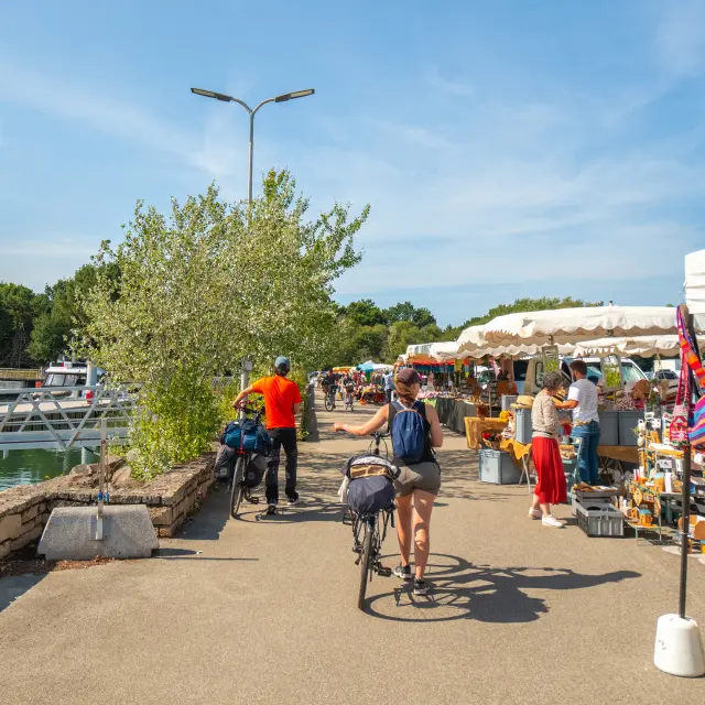 Une photo qui représente un marché en plein air le long d’un port, avec stands sous tentes blanches à droite, des personnes qui marchent, une personne à vélo au centre, et des bateaux/mâts à gauche près de l’eau, sous un ciel bleu.