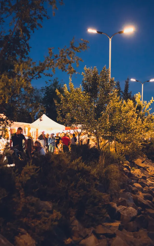 La photo montre une scène de soirée/nuit en extérieur, au bord de l’eau. Des lampadaires allumés éclairent une zone avec des arbres et des étales blanches de marchés, où un petit groupe de personnes se tient ou circule. Au premier plan, un talus rocheux et de la végétation. À droite, une marina ou des pontons avec des bateaux, et de l’eau sombre.