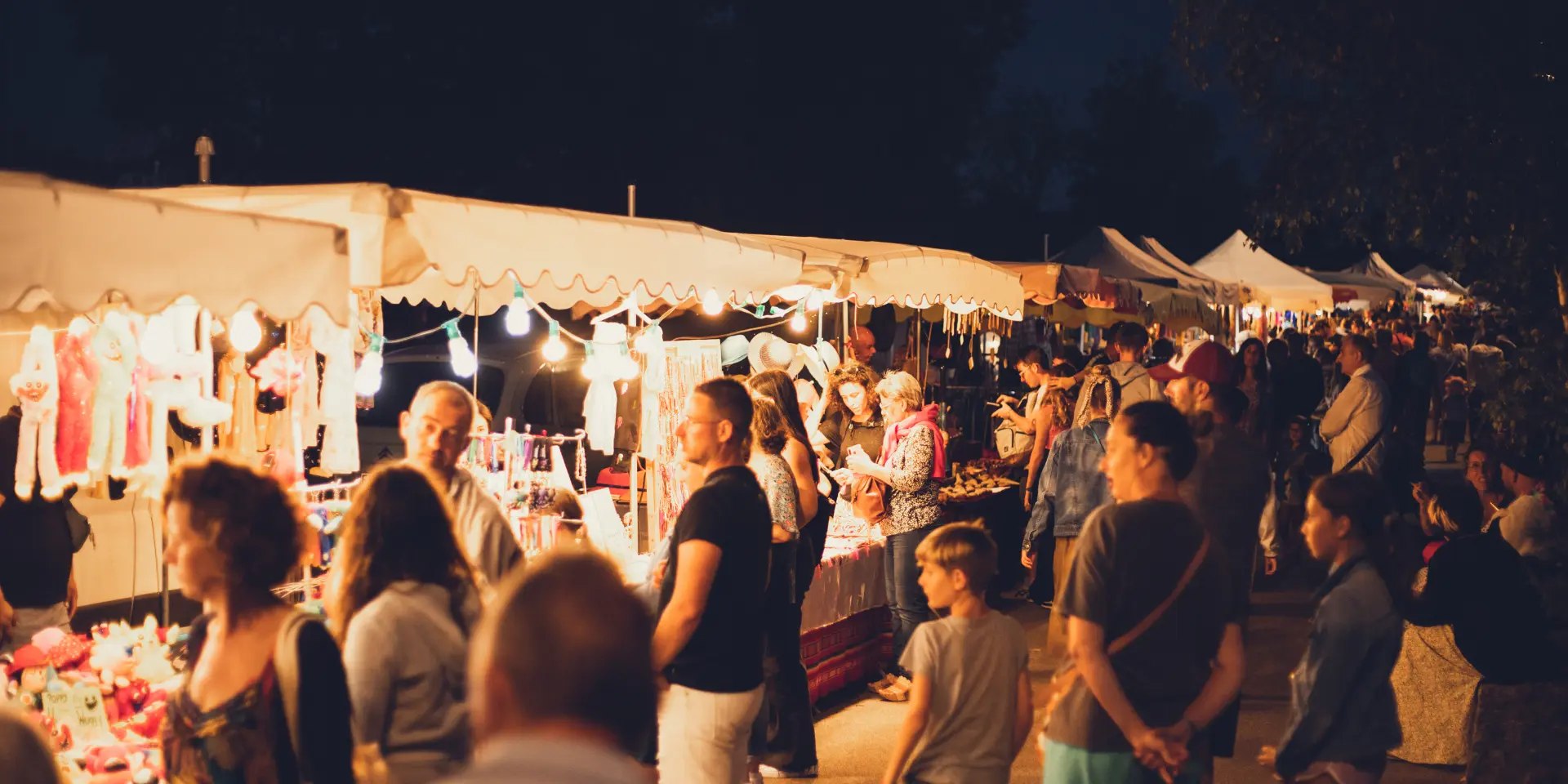 La photo montre un marché nocturne: une rangée de stands sous des tonnelles blanches éclairées de lumières chaudes. Plusieurs personnes (adultes et enfants) sont debout et circulent devant les étals. Des vêtements et divers articles pendent et sont exposés sur des présentoirs. L’arrière-plan est sombre (ciel de nuit) avec des silhouettes d’arbres; au premier plan, une zone floue de végétation.