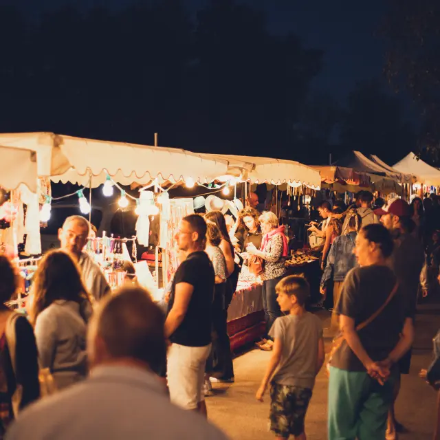 La photo montre un marché nocturne: une rangée de stands sous des tonnelles blanches éclairées de lumières chaudes. Plusieurs personnes (adultes et enfants) sont debout et circulent devant les étals. Des vêtements et divers articles pendent et sont exposés sur des présentoirs. L’arrière-plan est sombre (ciel de nuit) avec des silhouettes d’arbres; au premier plan, une zone floue de végétation.