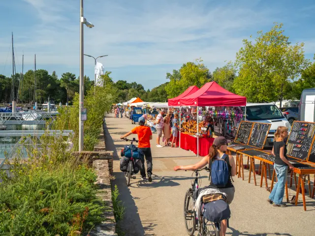 Dans la photo, une allée en bord de port avec des étals sous des tentes rouges et blanches. Des personnes se promènent, certaines à vélo. À gauche, des bateaux et mâts dans la marina. À droite, des panneaux/présentoirs de bijoux ou d’objets sont alignés. Ciel bleu, arbres verts au fond.