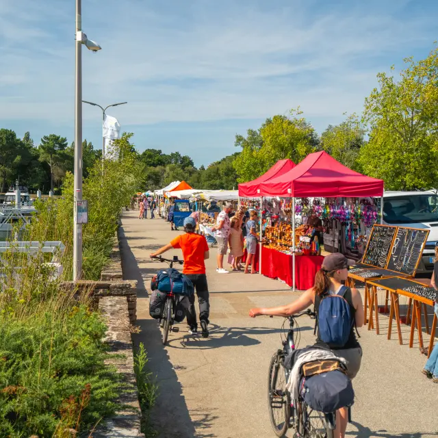Dans la photo, une allée en bord de port avec des étals sous des tentes rouges et blanches. Des personnes se promènent, certaines à vélo. À gauche, des bateaux et mâts dans la marina. À droite, des panneaux/présentoirs de bijoux ou d’objets sont alignés. Ciel bleu, arbres verts au fond.