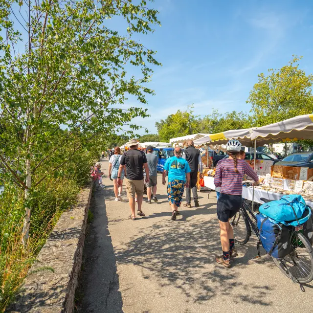 La photo montre une promenade ensoleillée le long d’un port de plaisance. À gauche, de l’eau avec des bateaux amarrés; au bord, un muret en pierre et des arbustes. Au centre, une allée piétonne avec plusieurs personnes qui marchent. À droite, des stands de marché sous des tentes blanches et jaunes, avec des tables couvertes de nappes blanches et des produits exposés. Un vélo chargé de sacs bleus est au premier plan côté droit. Ciel bleu, arbres verts.