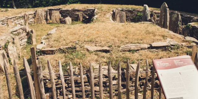 A photograph of a megalithic site. The photo shows a stone enclosure, a dry grassy area in the centre, upright slabs/menhirs, a small wooden fence in the foreground and an information panel in the lower right.