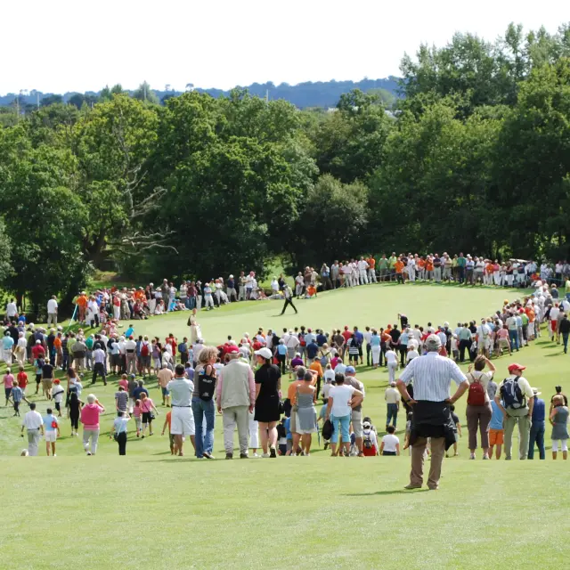 De foto toont een zacht glooiende groene fairway, omzoomd door bomen, met een grote menigte verzameld aan beide zijden rond een green of speelplaats; verschillende mensen lopen op het gras, en het tafereel ziet eruit als een evenement of toernooi.