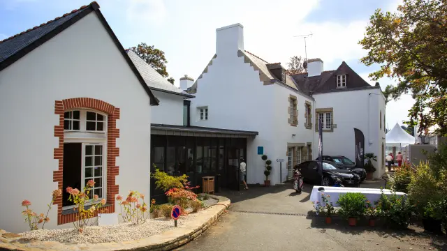 Une photo montre l’extérieur d’un bâtiment blanc de style manoir/club-house, avec encadrements de fenêtres en briques rouges sur une aile à gauche, une allée goudronnée au premier plan, des plantes en pots et un massif fleuri. À droite de l’allée, une voiture noire est stationnée près de l’entrée. Plus loin, deux oriflammes sombres et une tente blanche sont visibles, avec quelques personnes debout près de la tente.
