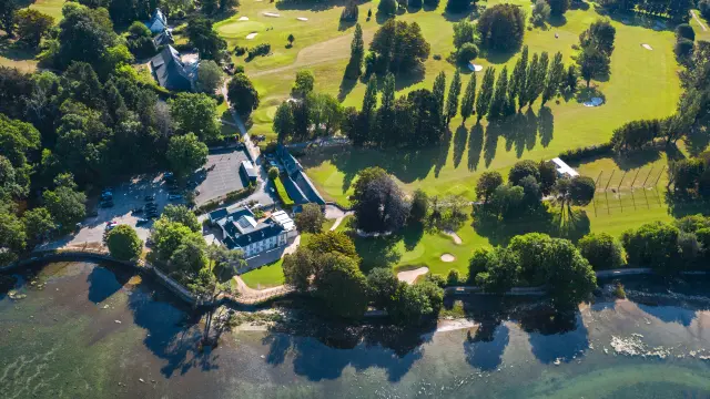 Une photo aérienne au centre-gauche montrant un golf en bord de mer: pelouses vertes avec greens et bunkers, des alignements d’arbres, un bâtiment type club-house, et de l’eau au premier plan.