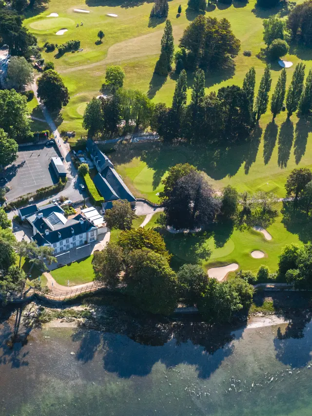 Une photo aérienne au centre-gauche montrant un golf en bord de mer: pelouses vertes avec greens et bunkers, des alignements d’arbres, un bâtiment type club-house, et de l’eau au premier plan.