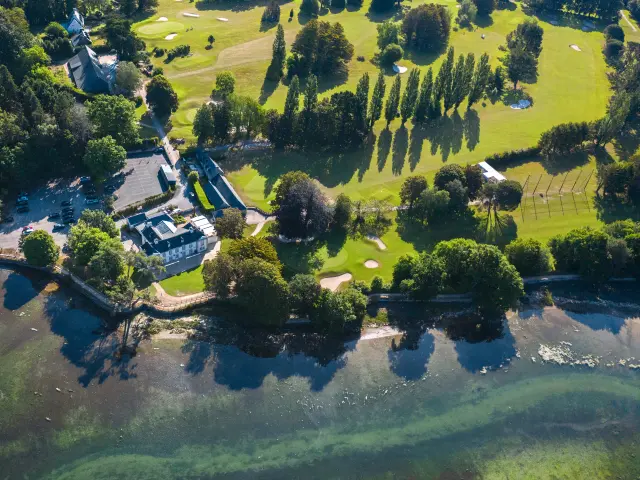 Ein Luftbild in der Mitte links zeigt einen Golfplatz am Meer: grüne Rasenflächen mit Greens und Bunkern, Baumreihen, ein Clubhaus und Wasser im Vordergrund.