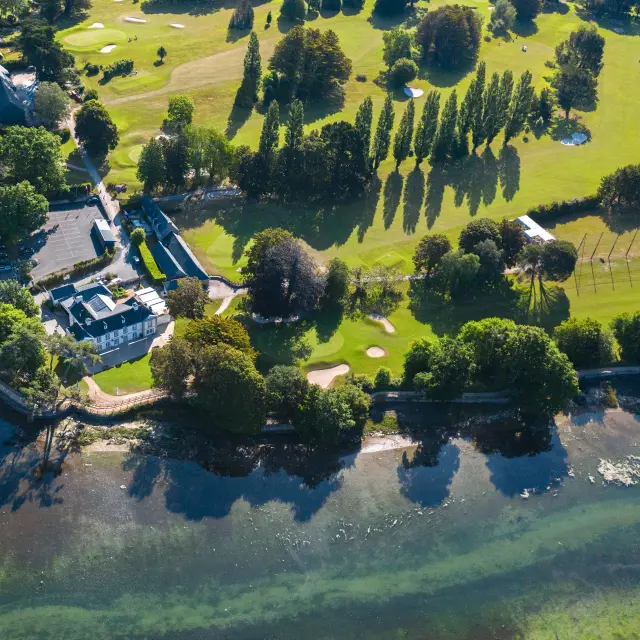 Ein Luftbild in der Mitte links zeigt einen Golfplatz am Meer: grüne Rasenflächen mit Greens und Bunkern, Baumreihen, ein Clubhaus und Wasser im Vordergrund.