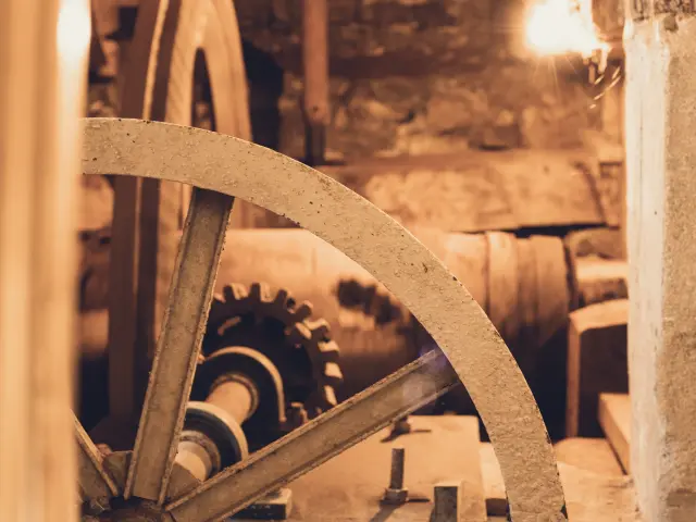 Das Foto zeigt einen Mühlenmechanismus in Sepiatönen, mit einem großen Metallrad im Vordergrund, Zahnrädern und einem Riemen/einer Achse im Hintergrund und einem großen Stein- oder Betonblock auf der rechten Seite.