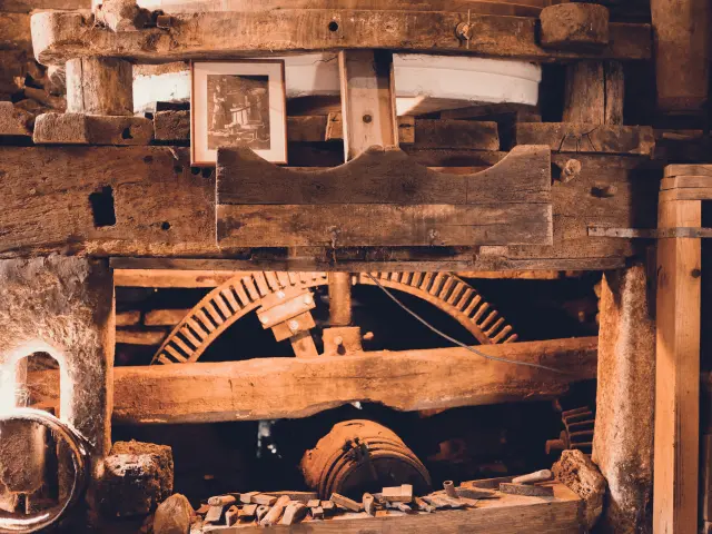 Photo d'un mécanisme ancien en bois, type moulin. Plusieurs poutres épaisses et usées, une grande roue dentée en bois partiellement visible au centre, des pièces métalliques et outils en bas, et une petite photo encadrée posée en haut sur la structure. Tons brun/orangé, ambiance d’atelier ancien.