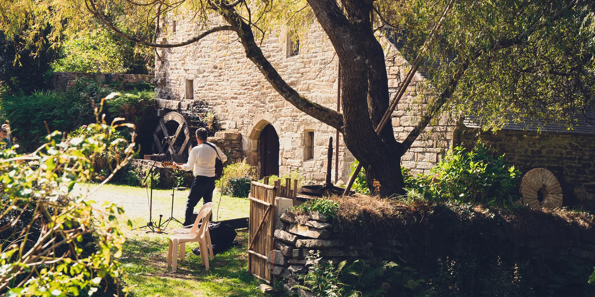 Une photo qui montre un vieux moulin en pierre dans un jardin boisé, avec un grand arbre au premier plan et une roue de moulin visible près du bâtiment; une personne se tient dans l’herbe près d’une petite barrière et de chaises.