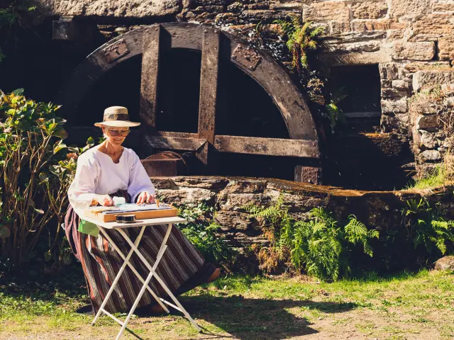 La photo montre une grande roue de moulin en bois contre un mur en pierre. Au premier plan, une femme âgée à peau claire, portant un chapeau de paille et un haut blanc, est assise devant une petite table pliante, en train de peindre ou dessiner. Végétation (fougères/herbes) au sol autour.
