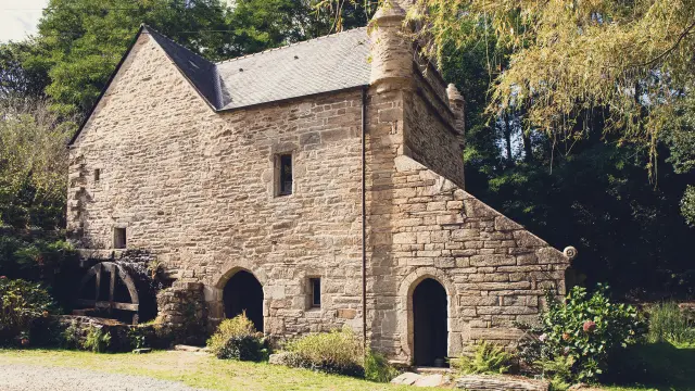 A photo of an old stone mill. The grey stone building has a slate roof, a large arched door on the ground floor, a small window in the middle, and a mill wheel visible on the left. Trees and vegetation surround the building; gravel and grass are in the foreground.