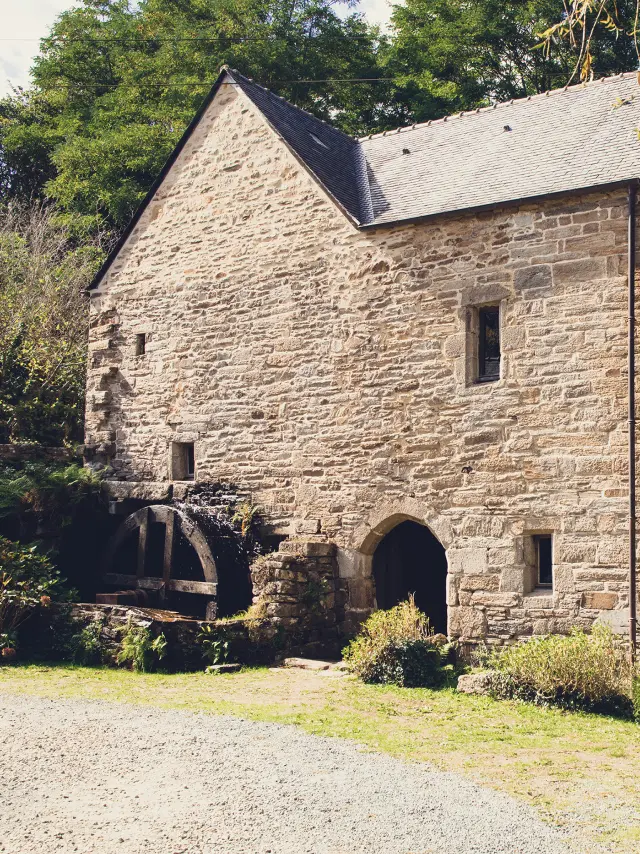 Une photo d’un vieux moulin en pierre. Le bâtiment en pierre grise a un toit en ardoise, une grande porte en arc au rez-de-chaussée, une petite fenêtre au milieu, et une roue de moulin visible sur la gauche. Des arbres et de la végétation entourent le bâtiment; du gravier et de l’herbe sont au premier plan.