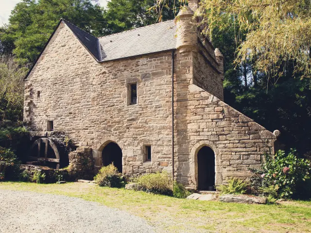 Ein Foto von einer alten Steinmühle. Das Gebäude aus grauem Stein hat ein Schieferdach, eine große Bogentür im Erdgeschoss, ein kleines Fenster in der Mitte und ein Mühlrad, das auf der linken Seite zu sehen ist. Bäume und Vegetation umgeben das Gebäude; Kies und Gras sind im Vordergrund.