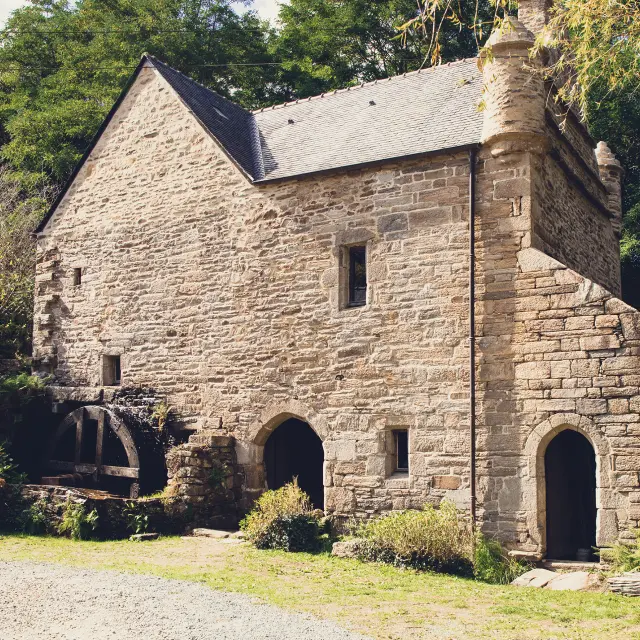 Ein Foto von einer alten Steinmühle. Das Gebäude aus grauem Stein hat ein Schieferdach, eine große Bogentür im Erdgeschoss, ein kleines Fenster in der Mitte und ein Mühlrad, das auf der linken Seite zu sehen ist. Bäume und Vegetation umgeben das Gebäude; Kies und Gras sind im Vordergrund.