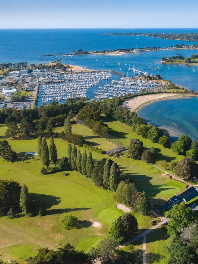 Photo showing a coastline with a wide bay/estuary, a harbour with numerous boats on the left, green golf areas in the foreground and dwellings on the right, then the sea in the background.
