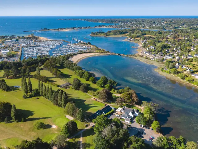 Foto, das eine Küste mit einer breiten Bucht/Mündung zeigt, einen Hafen mit vielen Booten auf der linken Seite, grüne Golfplätze im Vordergrund und Wohnhäuser auf der rechten Seite, dann das Meer im Hintergrund.